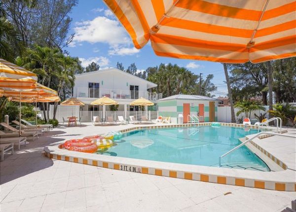 Outdoor pool area with yellow-orange striped umbrellas, lounge chairs, and palm trees, pastel buildings in the background on a sunny day.